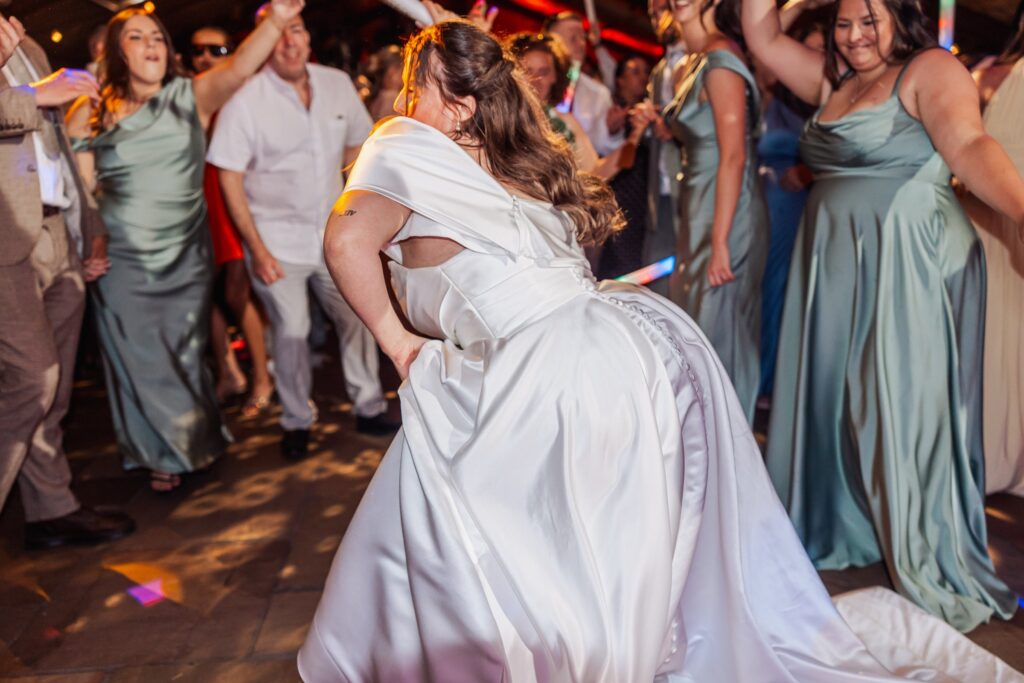 A woman in a white wedding dress dances on a lively wedding reception floor surrounded by guests in formal attire
