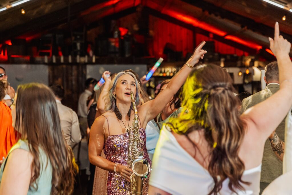 A woman in a sequined dress dances with a saxophone among guests at a wedding reception, with red lighting and sparklers in the air at Autumn & Sam - Owen house barn
