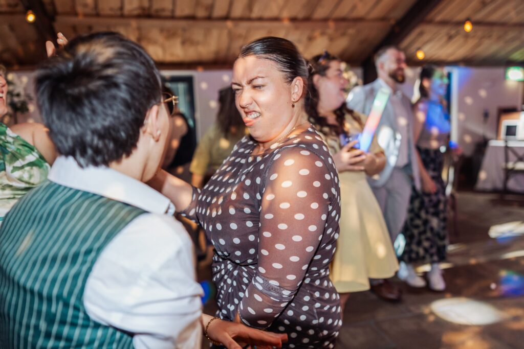 Two people are dancing closely together in a warmly lit wooden room with fairy lights, surrounded by other guests at a wedding reception
