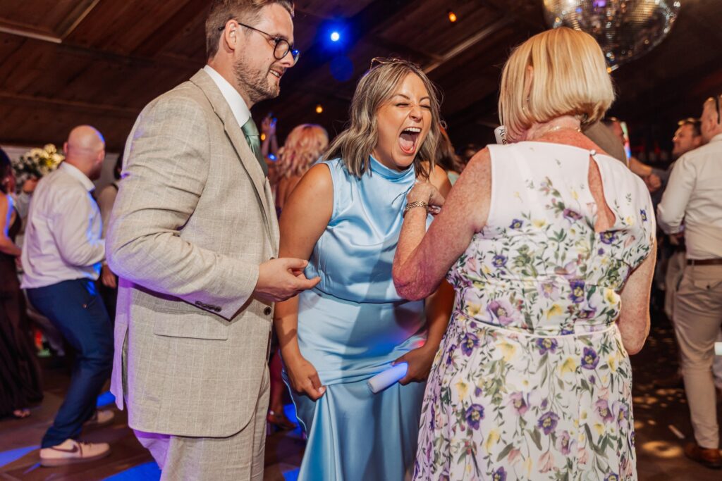 A woman in a light blue dress laughs and interacts with another woman in a floral dress at a wedding reception with wooden at Autumn & Sam - Owen house barn