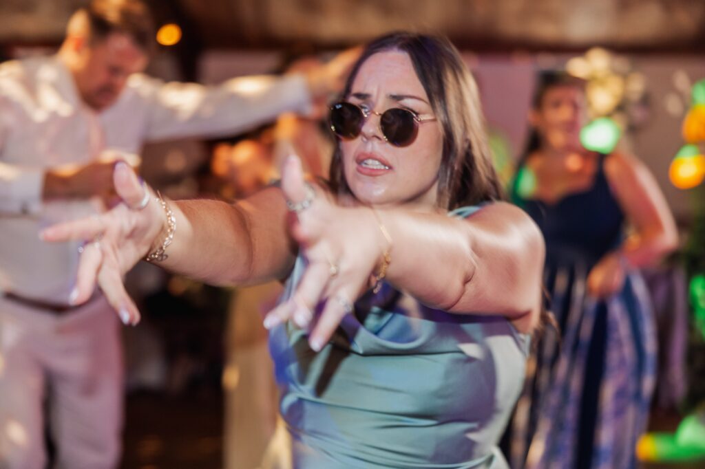 A woman in a teal dress dances energetically, wearing sunglasses and jewellery at Autumn & Sam - Owen house barn