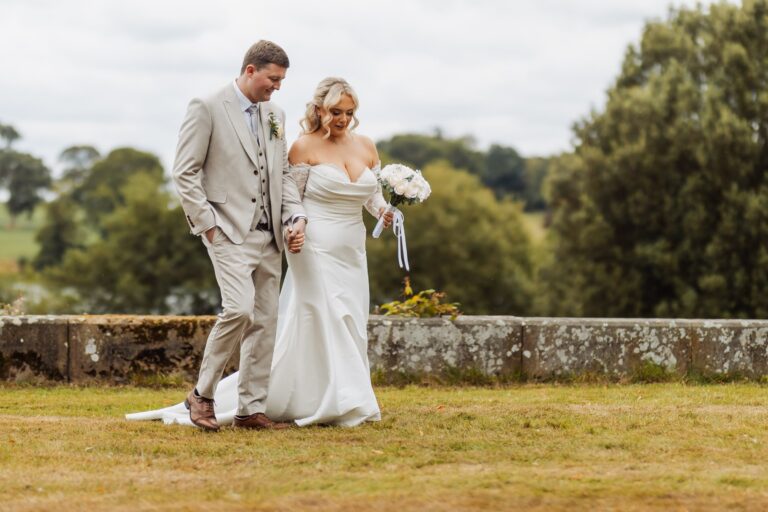 A couple in wedding attire walks hand in hand on a grassy area, with the bride holding a bouquet of white flowers at Coniston Hotel, Skipton