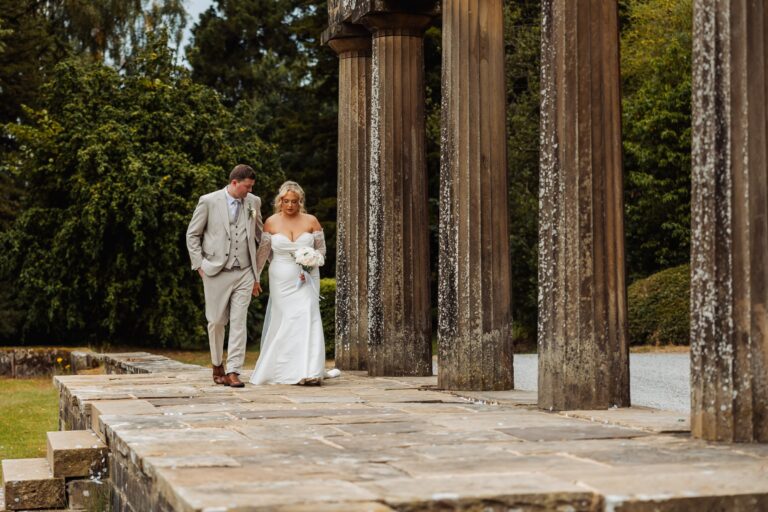 A couple in wedding attire walks together on a stone pathway lined with tall, weathered columns