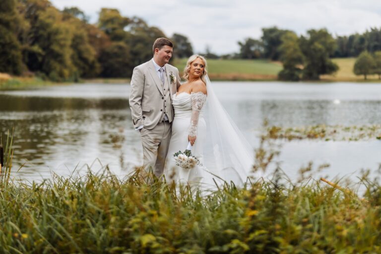 A couple in wedding attire stands close together by a lake, with the bride holding a bouquet of white flowers at Coniston Hotel, Skipton