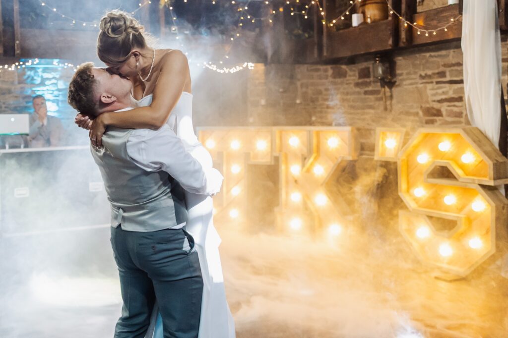 A couple in wedding attire embraces and kisses, surrounded by a smoky atmosphere and illuminated 'LOVE' letters, with fairy lights in the background