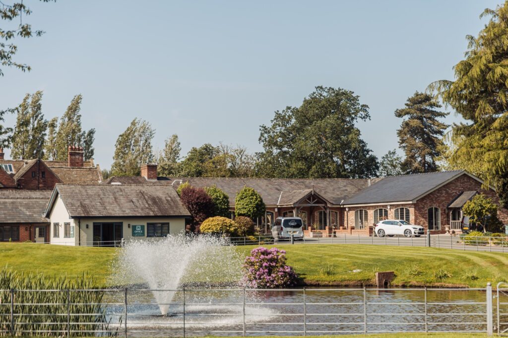 A fountain sprays water in the foreground, surrounded by lush greenery and traditional brick buildings under bright sunlight at Colshaw Hall, Cheshire