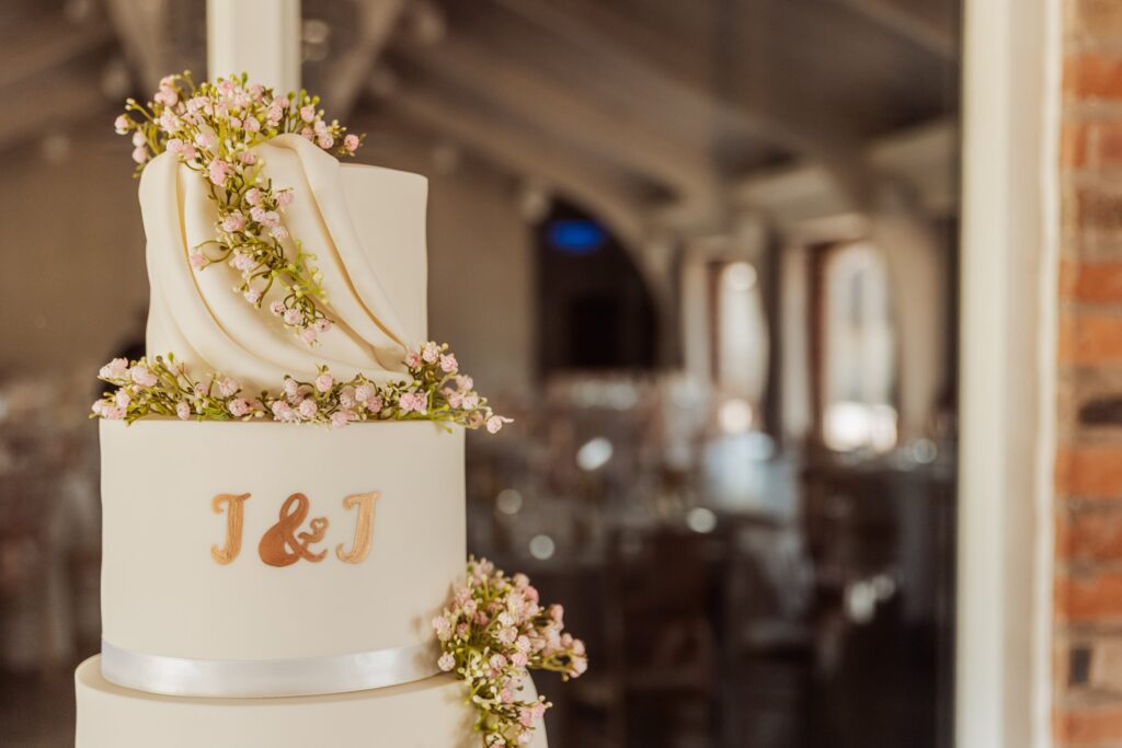 A tiered wedding cake with white fondant and gold lettering spelling 'J & J', decorated with pink flowers and a draped white ribbon at Colshaw Hall, Cheshire