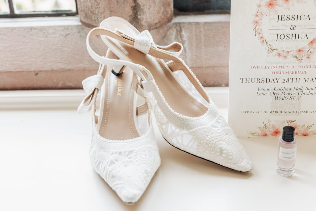 A pair of white lace wedding shoes with ankle straps, a perfume bottle labelled 'Purple Oud', and a wedding invitation for Jessica and Joshua