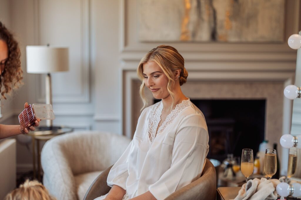A woman in a white lace robe is seated with another person holding a makeup palette nearby at Colshaw Hall, Cheshire