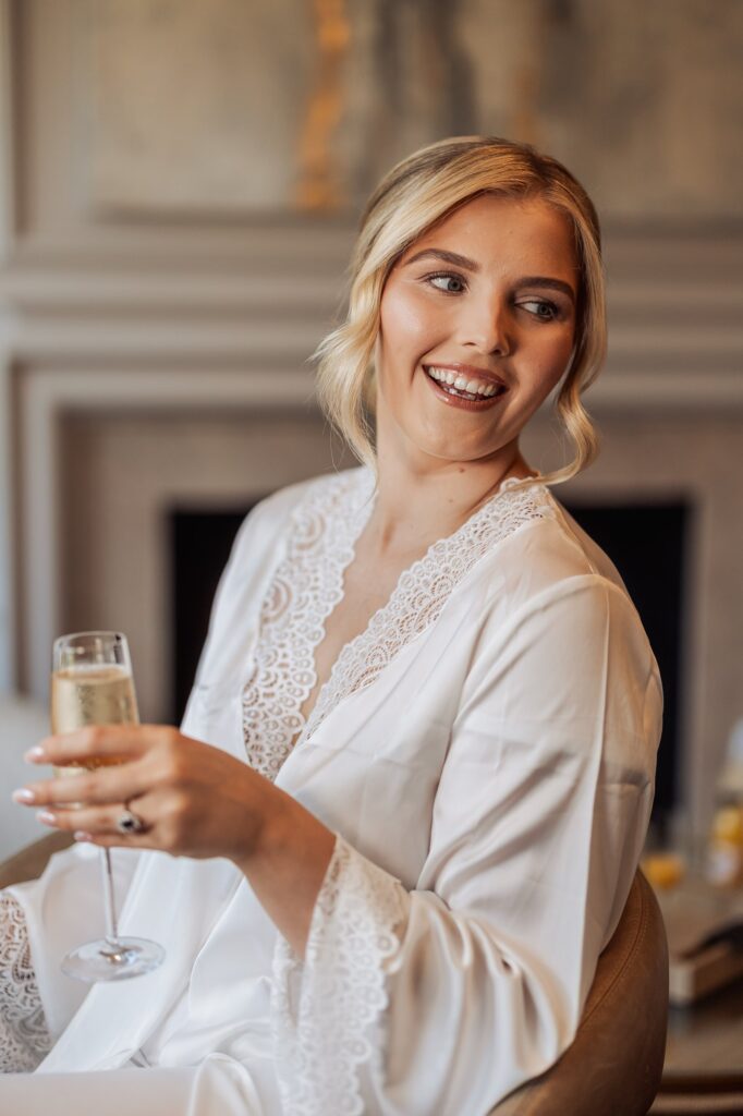 A woman in a white lace robe holds a glass of champagne, smiling and looking to the side at Colshaw Hall, Cheshire