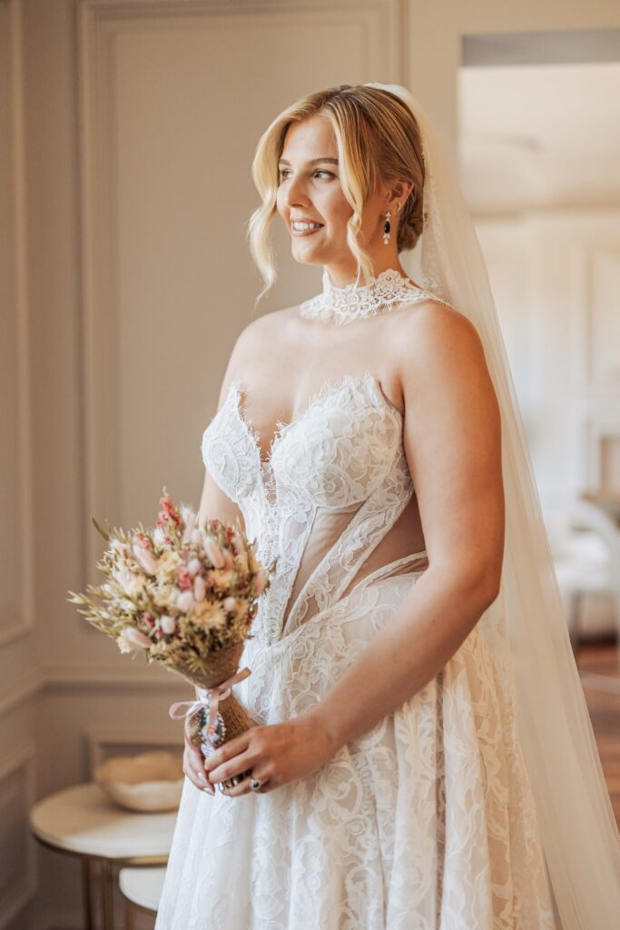 A woman in a white lace wedding dress holds a bouquet of dried flowers, standing indoors with a wooden floor and a door in the background