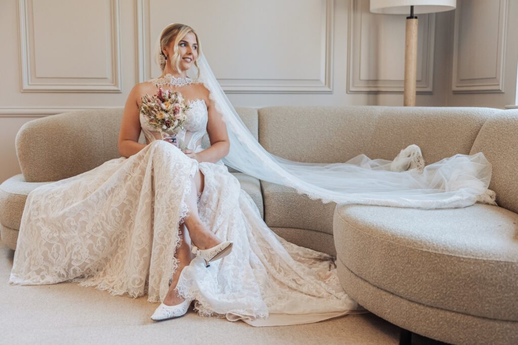 A bride in a lace wedding dress sits on a beige sofa, holding a bouquet of flowers, preparing for her wedding at Colshaw Hall, Cheshire