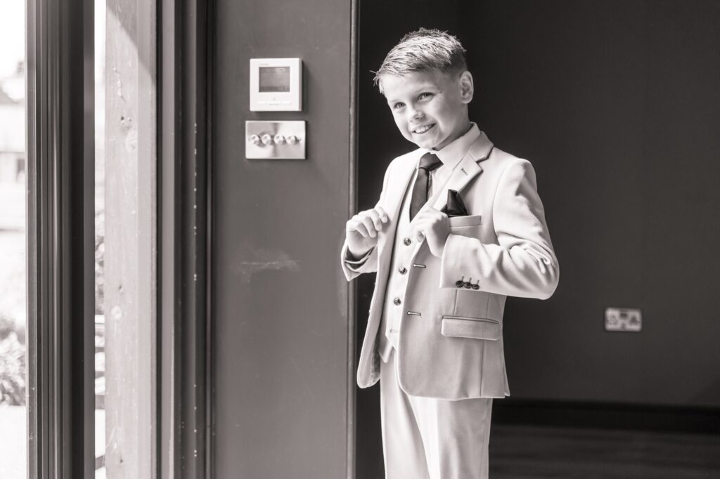 A young boy in a formal suit and tie adjusts his jacket in front of a dark wall with a thermostat and light switches