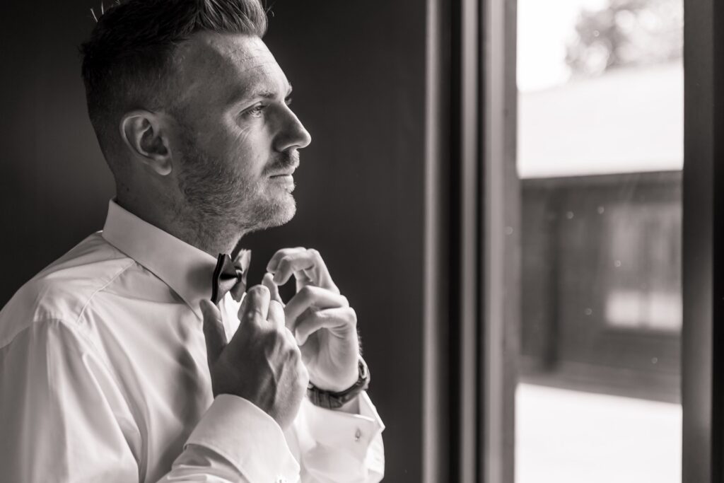 A man in a white shirt and bow tie adjusts his bow tie in front of a window, wearing a watch on his left wrist at Colshaw Hall, Cheshire