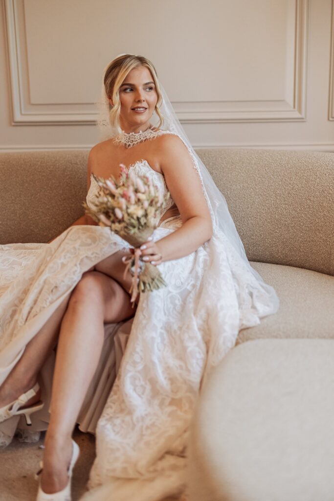 A bride in a lace wedding dress sits on a beige sofa, holding a bouquet of dried flowers, preparing for her wedding