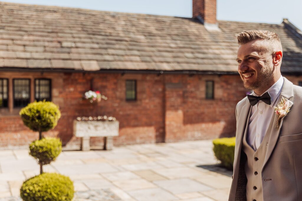 A man in a light grey suit with a black bow tie and a boutonniere stands in front of a brick building with a tiled roof at Colshaw Hall, Cheshire
