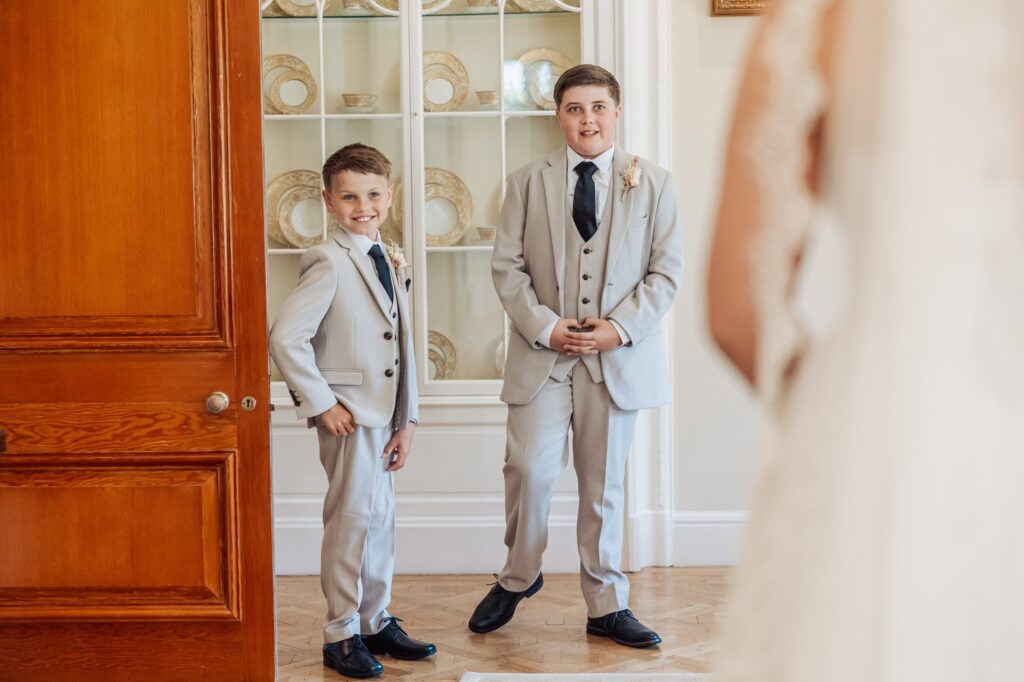 Two young boys in matching light grey suits and black ties stand in a warmly lit room at Colshaw Hall, Cheshire