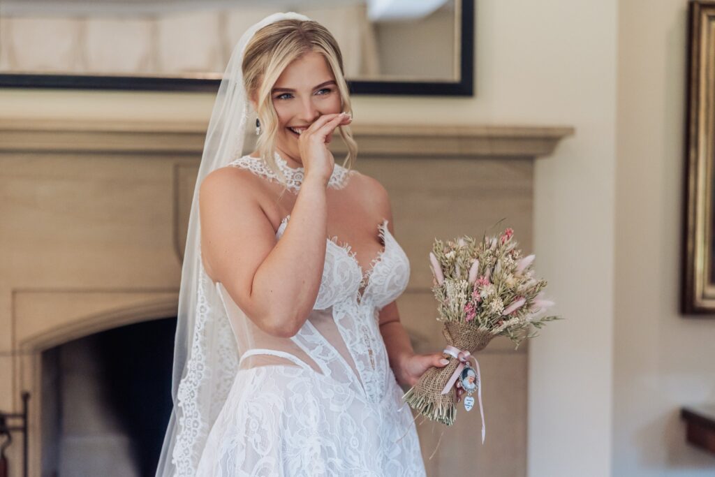 A bride in a lace wedding dress holds a bouquet of dried flowers and smiles, with her hand near her mouth, in a warmly lit room at Colshaw Hall, Cheshire