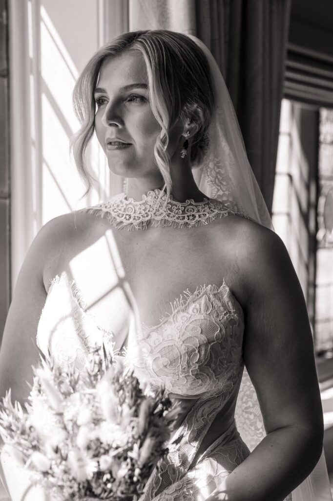 A bride in a lace wedding dress holds a bouquet of flowers, standing indoors with natural light coming through a window at Colshaw Hall, Cheshire