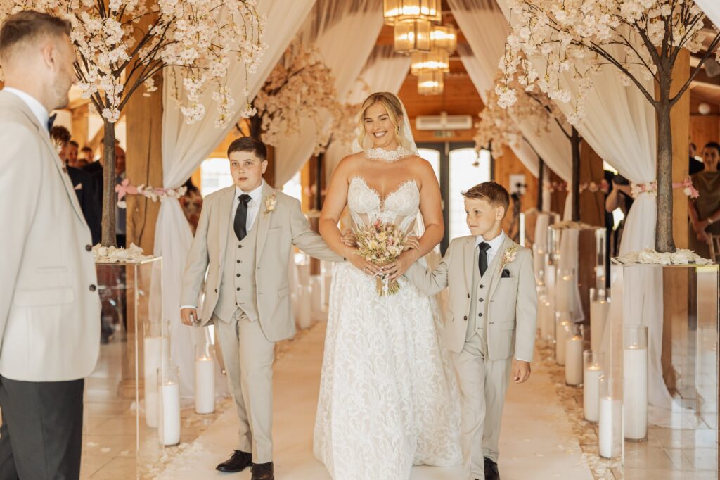 A bride in a lace wedding dress walks down an aisle flanked by two young boys in matching suits at Colshaw Hall, Cheshire
