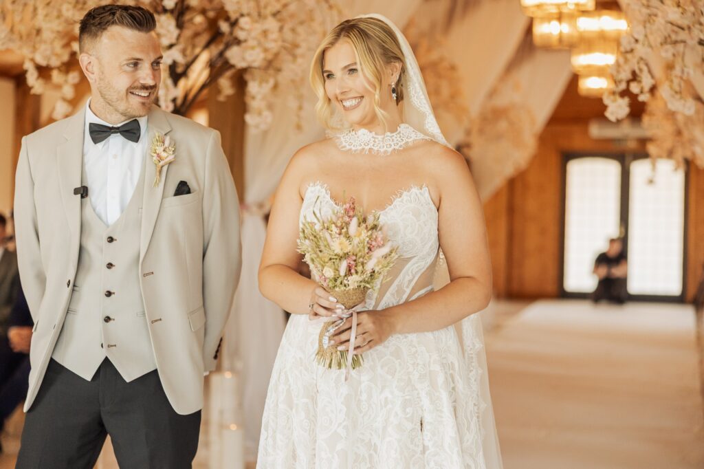 A couple in wedding attire stands in a warmly lit wooden hall, the bride holding a bouquet of dried flowers and wearing a lace wedding dress with a veil