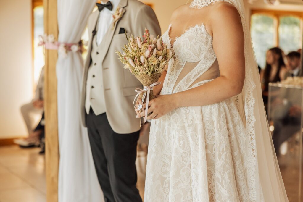 A bride in a lace wedding dress holds a bouquet of dried flowers, standing in a warmly lit room with wooden flooring and large windows at Colshaw Hall, Cheshire