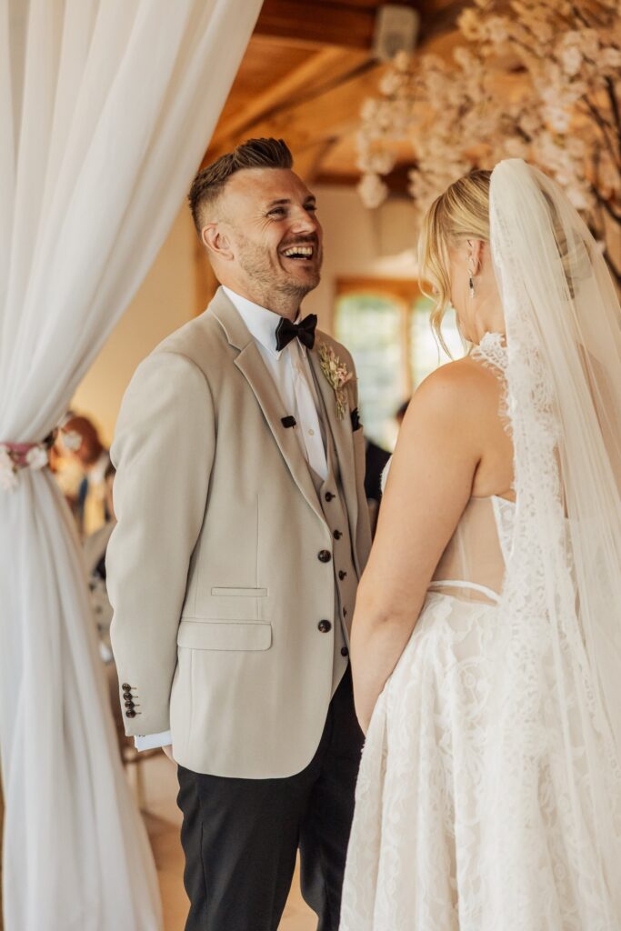A couple in wedding attire stands close together in a warmly lit wooden hall, facing each other during a wedding ceremony at Colshaw Hall, Cheshire