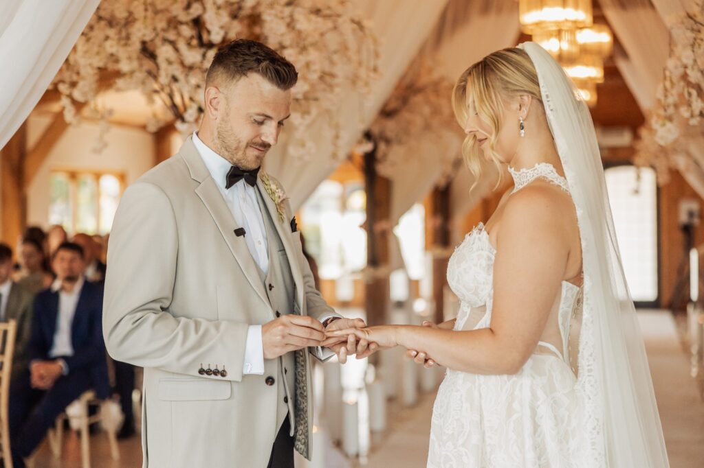 A couple exchanging wedding rings in a warmly lit wooden hall with floral decorations, surrounded by seated guests