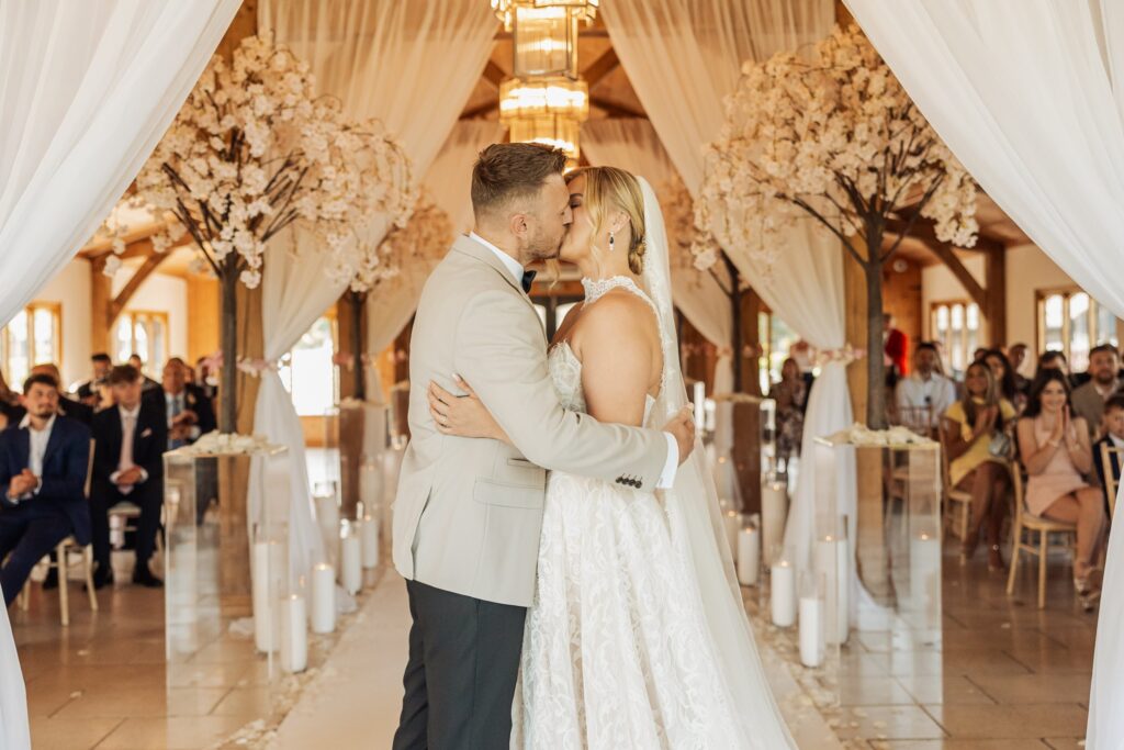 A couple in wedding attire share a kiss during a wedding ceremony in a warmly lit wooden hall with white drapes and decorative trees with white blossoms