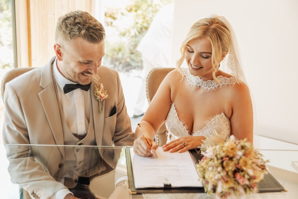 A couple in wedding attire signs the register at a table, with the bride in a lace dress and veil and the groom in a beige suit with a bow tie