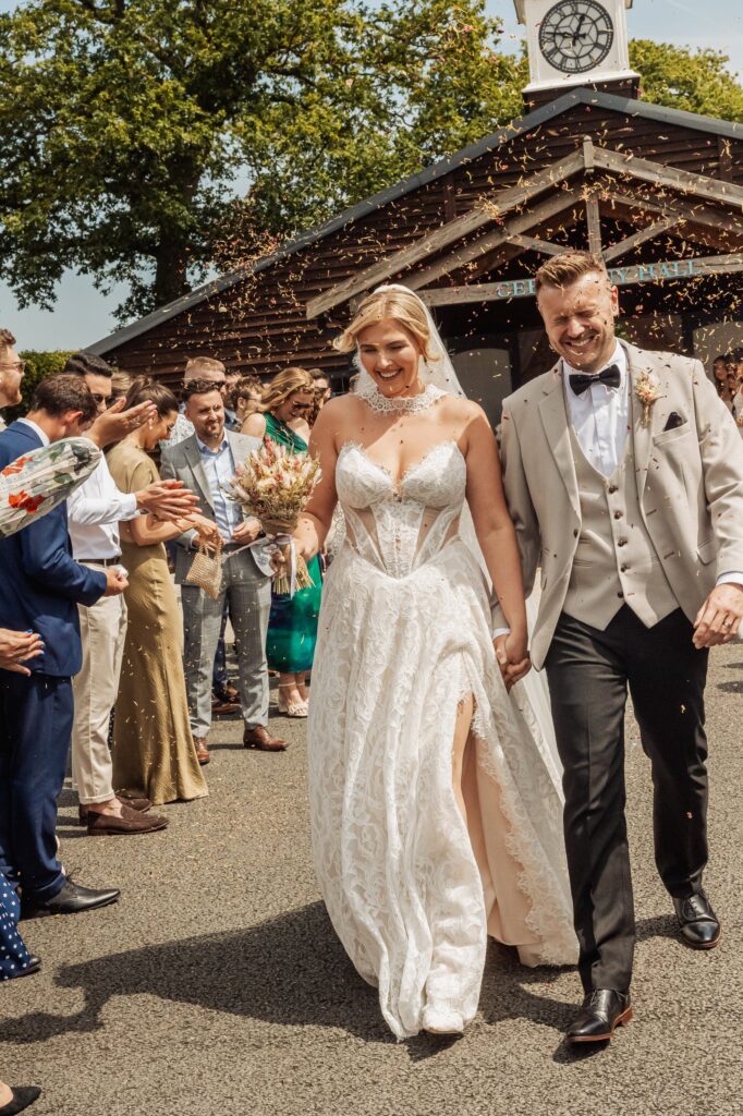 Confetti falls as a couple in wedding attire walks hand in hand, surrounded by guests in formal attire at Colshaw Hall, Cheshire