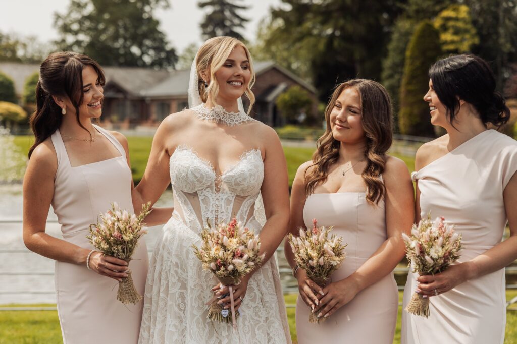 A bride in a lace wedding dress stands with three bridesmaids in matching pale pink dresses, holding bouquets of dried flowers at Colshaw Hall, Cheshire