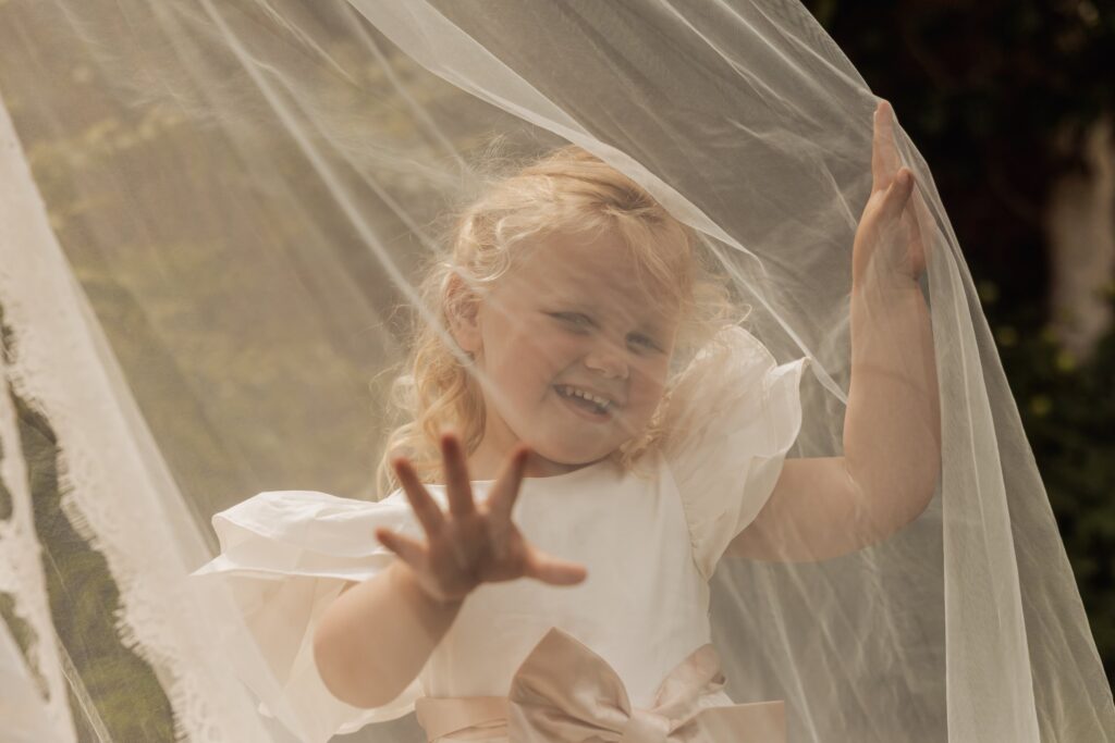 A young child in a white dress with a pink bow, holding a sheer white veil, smiling and waving