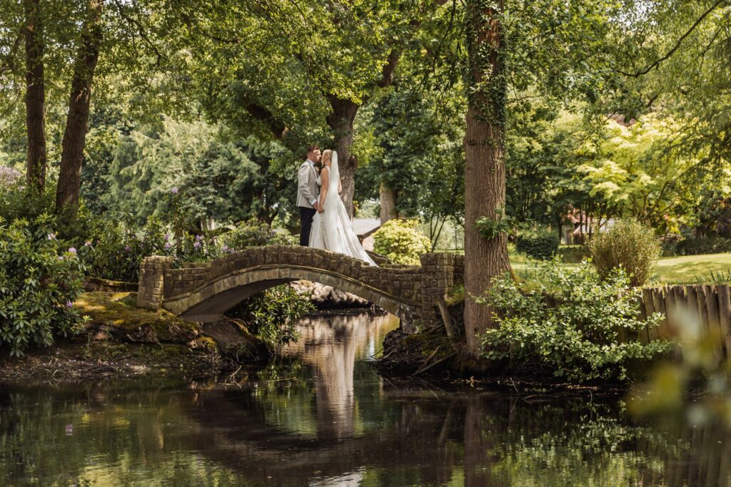 A couple in wedding attire stands on a stone bridge over a calm body of water, surrounded by lush greenery and trees at Colshaw Hall, Cheshire