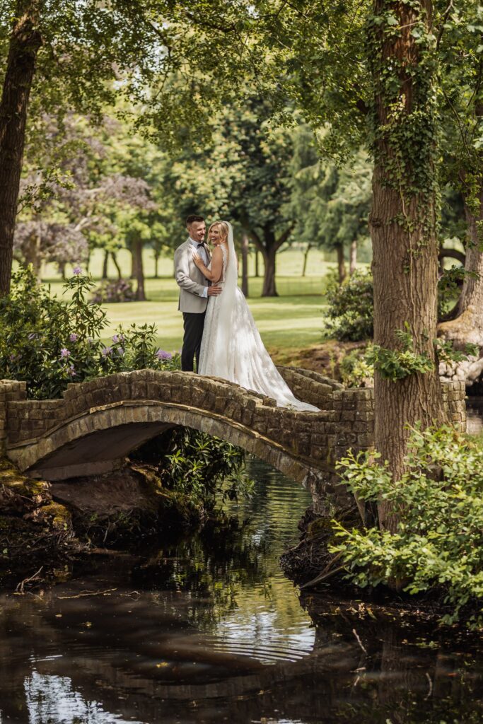 A couple in wedding attire stands on a stone bridge over a calm stream, surrounded by lush greenery and trees at Colshaw Hall, Cheshire