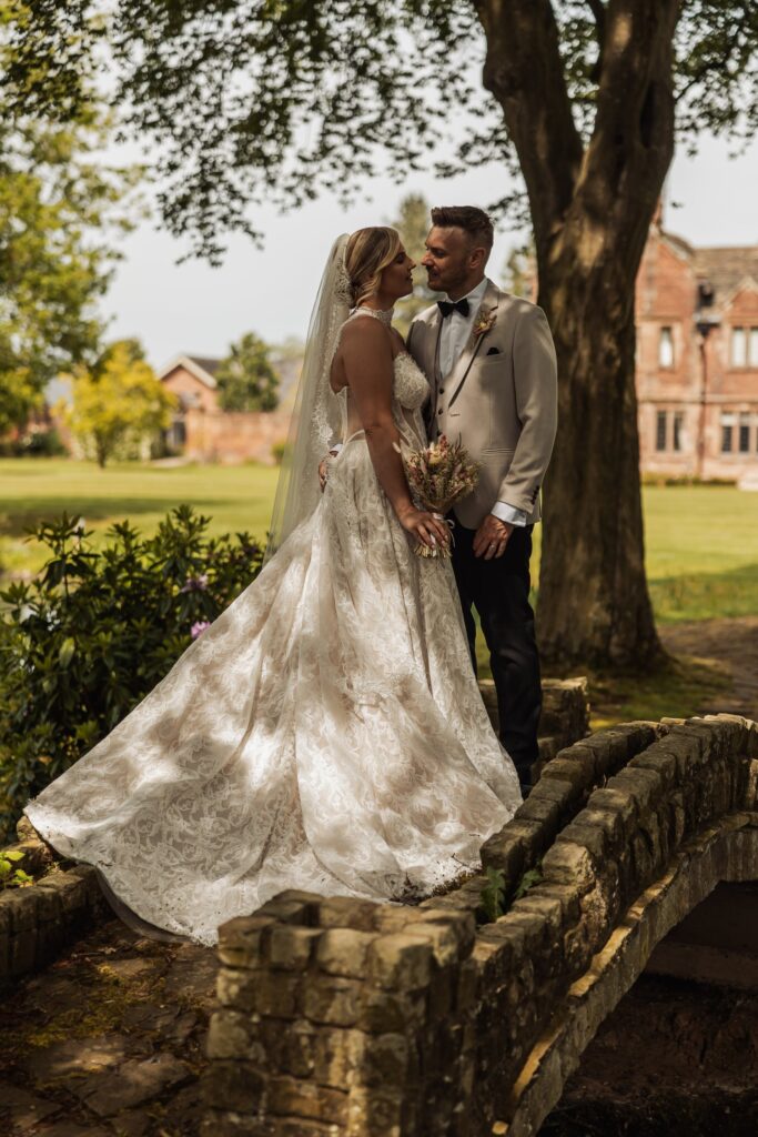 A couple in wedding attire stands close together on a stone bridge, with the bride in a lace gown and the groom in a light-coloured suit