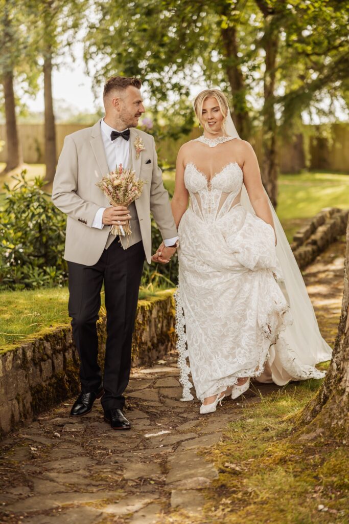 A couple in wedding attire walks hand in hand on a stone path in a garden at Colshaw Hall, Cheshire