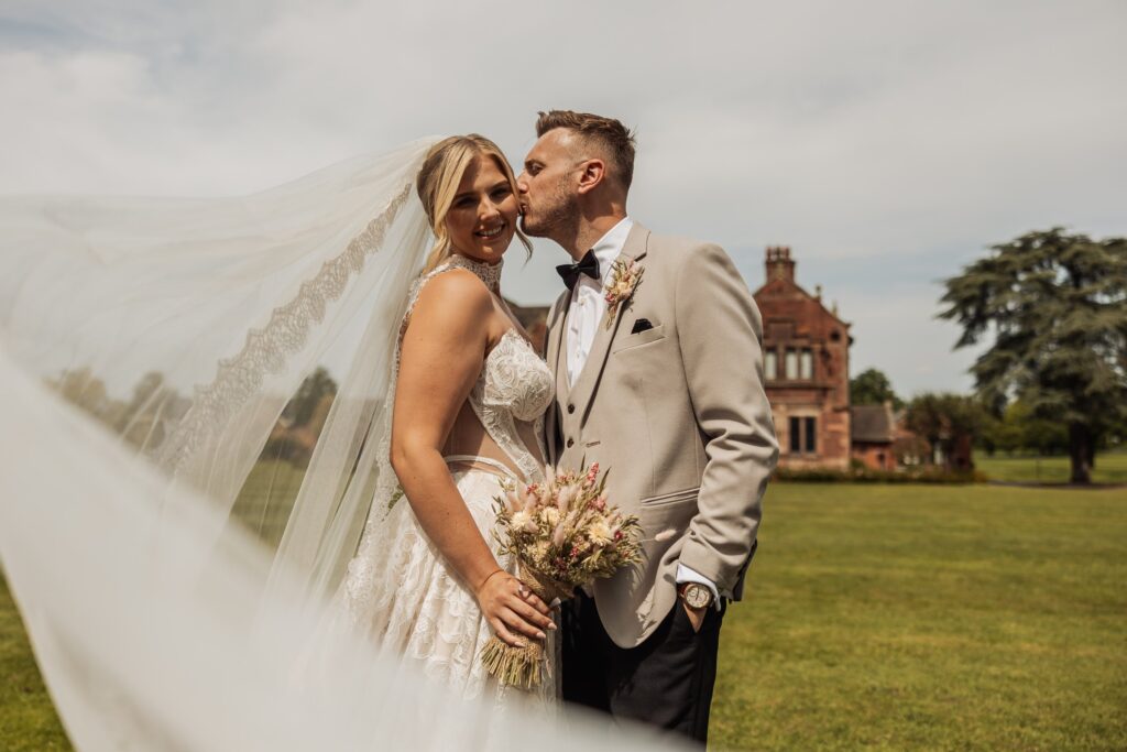 A couple in wedding attire stands close together outdoors, with the bride holding a bouquet and the groom in a beige suit