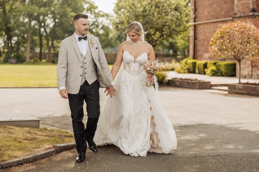 A couple in wedding attire walks hand in hand on a paved path with a grassy area and trees in the background