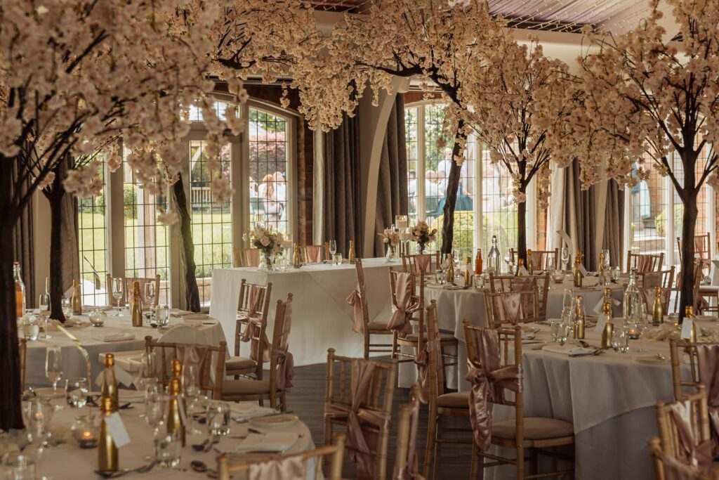 A warmly lit room with wooden flooring and large windows, decorated with artificial cherry blossom trees and white tablecloths, set up for a wedding reception