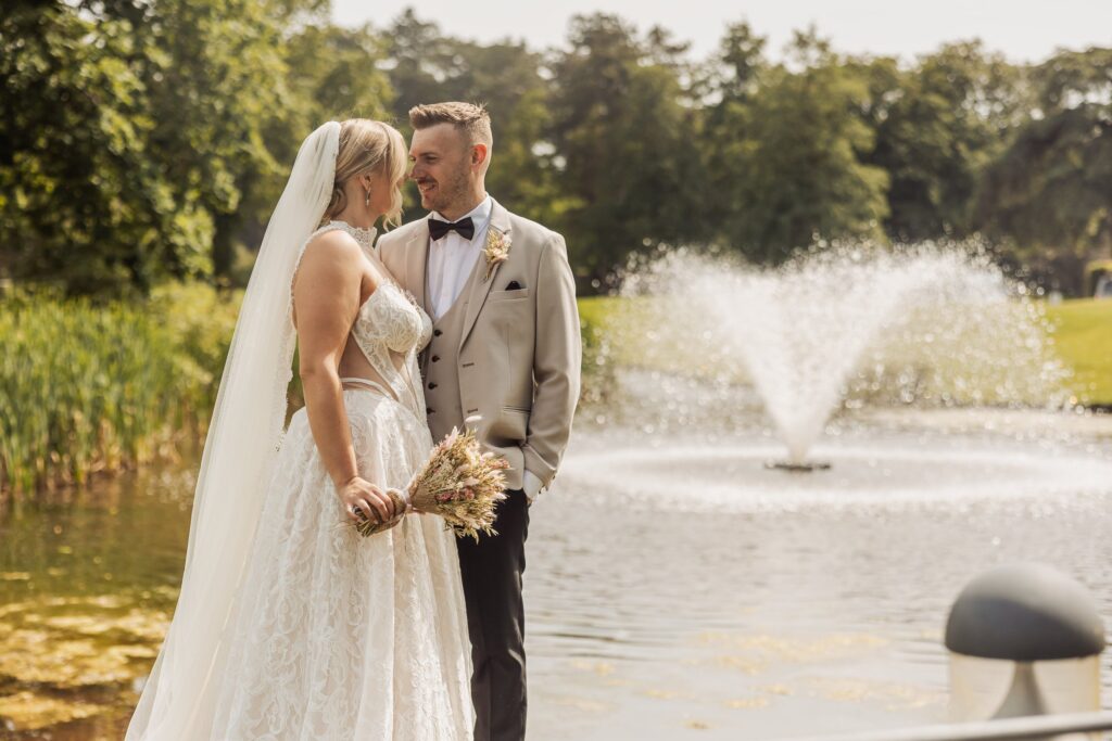 A couple in wedding attire stands close together by a fountain, with the bride holding a bouquet and the groom in a beige suit with a bow at Colshaw Hall, Cheshire