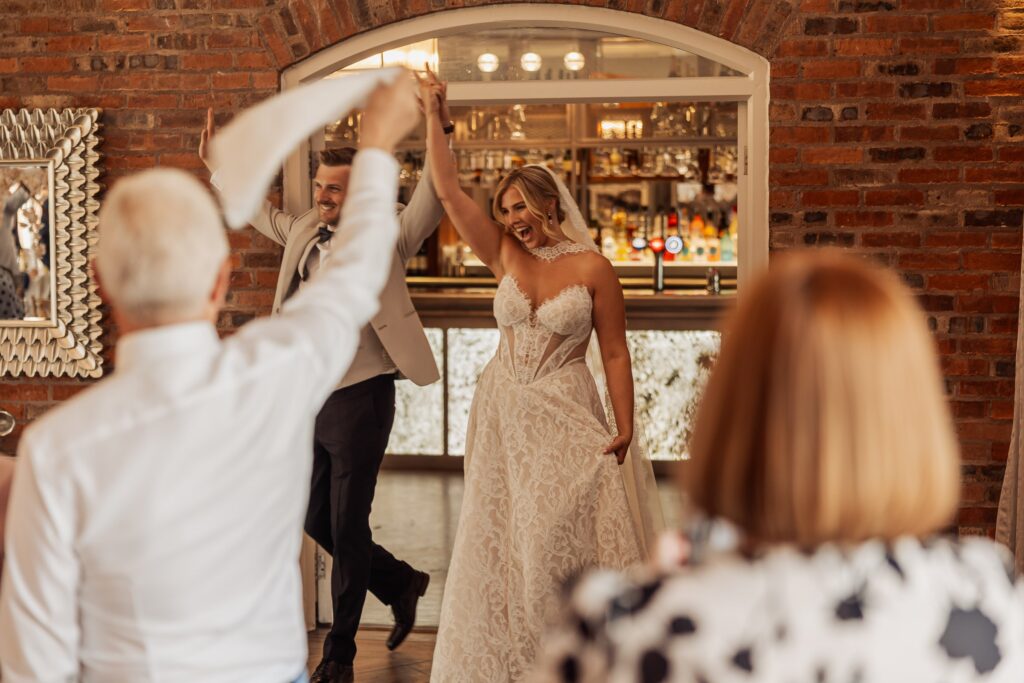 A couple in wedding attire dances mid-movement on a wooden floor, with guests watching from the edges in a warmly lit room with a brick at Colshaw Hall, Cheshire