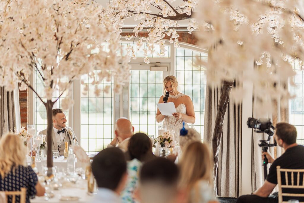 A bride in a white lace dress stands at a podium, holding a microphone and reading from a piece of paper, addressing a seated audience at a wedding reception