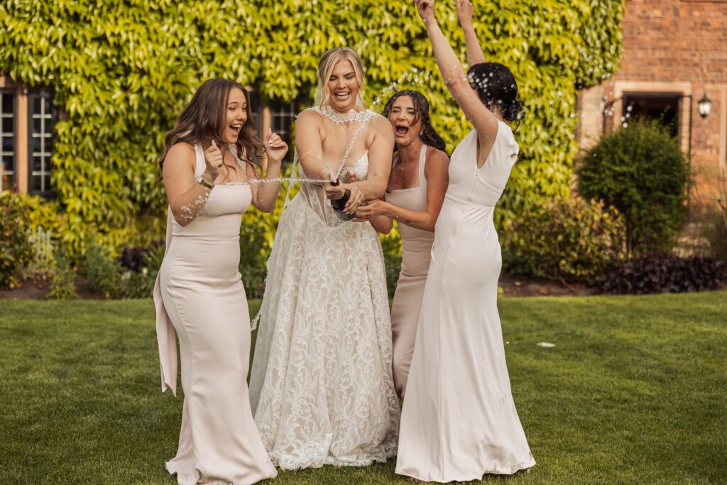 A bride in a lace wedding dress is surrounded by three bridesmaids in pale pink dresses, celebrating with champagne on a grassy lawn at Colshaw Hall, Cheshire