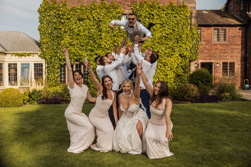 A group of bridesmaids in matching dresses lift a man in a suit and bow tie into the air on a grassy lawn at Colshaw Hall, Cheshire
