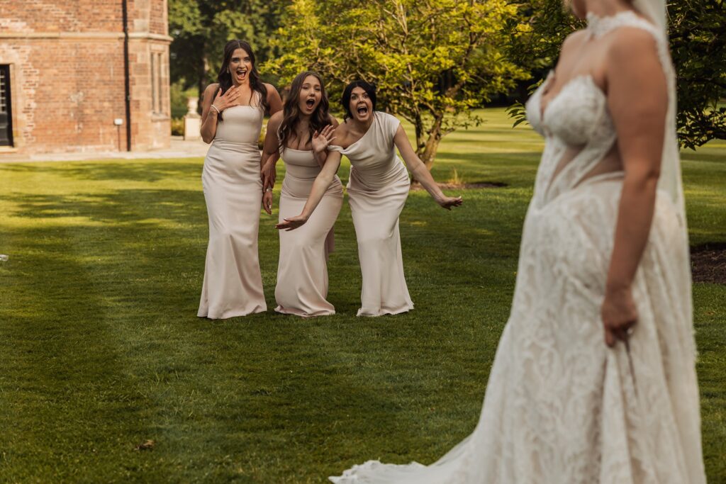 Three bridesmaids in matching dresses react with surprise and excitement as they see the bride in her white lace wedding gown on a grassy at Colshaw Hall, Cheshire