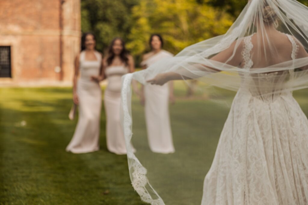 A bride in a lace wedding dress holds her veil, with three bridesmaids in matching white dresses standing behind her on a grassy area at Colshaw Hall, Cheshire