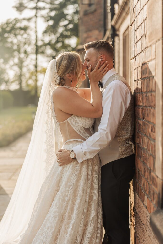 A couple in wedding attire stands close together, embracing, against a brick wall outdoors with trees and sunlight in the background at Colshaw Hall, Cheshire