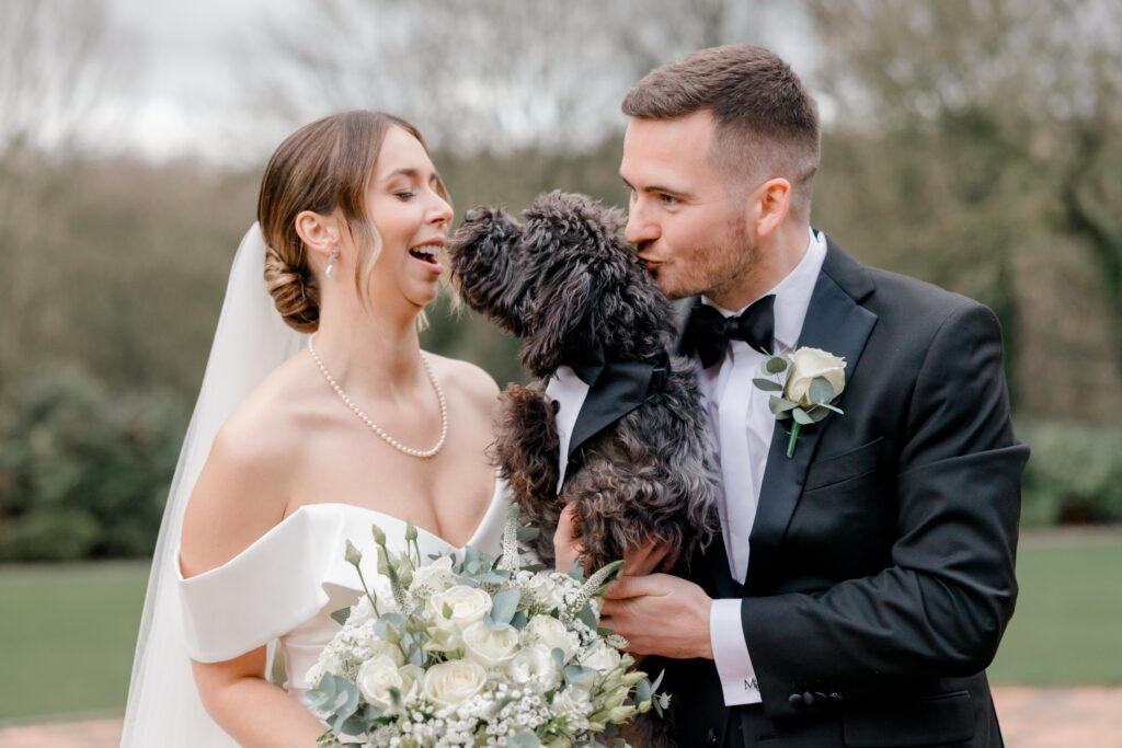 Bride laughing as the groom kisses their black cockapoo, who is wearing a tuxedo collar