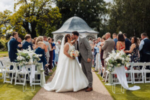 Bride and groom kiss at the end of the aisle during their outdoor ceremony at Hodsock Priory, guests standing either side